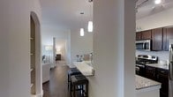 This well-appointed single-floor residence showcases a thoughtfully designed open-concept layout that seamlessly connects living and dining areas. The interior is anchored by rich, dark hardwood flooring that flows throughout, creating a warm and cohesive aesthetic against the clean white walls and architectural archways.

A standout feature of the space is the granite-topped breakfast bar, which extends from the kitchen and is illuminated by two elegant cylindrical pendant lights that hang at varying heights, adding visual interest and a modern touch. The bar is paired with upholstered counter stools accented with nailhead trim, offering both style and functionality as a casual dining or entertaining area.

The kitchen is equipped with a sleek modern faucet and ample counter space, reflecting a practical yet refined design sensibility. The open hallway leads to private bedroom spaces, which are visible in the background and appear well-lit with warm, ambient lighting, suggesting comfortable and inviting retreats. The overall ambiance of the residence is contemporary and polished, with a neutral color palette that provides a versatile backdrop for a variety of furnishing styles. The layout offers a smooth, natural flow between spaces, making the home feel both spacious and welcoming.