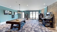Located at 1800 Indigo Square in Mount Pleasant, South Carolina, this single-floor community recreation space is designed for socializing and entertainment. The game room is thoughtfully appointed with a full-size pool table positioned beneath a trio of stylish industrial pendant lights, creating a focused and inviting atmosphere for play. An upright arcade cabinet stationed along one wall adds a fun, retro element to the space, accompanied by a bar-height stool for casual seating. The room features teal-painted walls that lend a modern, energetic feel, complemented by an exposed brick accent wall that adds warmth and texture. A bold geometric-patterned carpet in shades of blue and gray grounds the space with a contemporary aesthetic. Large multi-pane windows and glass-paneled doors flood the interior with natural light while offering views of the surrounding greenery outside. A small seating nook near the windows provides a relaxed spot to unwind between games. Recessed ceiling lighting and crown molding complete the polished look. A wall-mounted television adds a multimedia element, making this space well-suited for gatherings, casual hangouts, and community events alike.