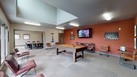 Located at 20917 44th Ave W in Lynnwood, Washington, this single-floor community amenity space is thoughtfully designed to serve both recreational and professional needs. The layout is divided into distinct functional zones that flow naturally into one another.

The centerpiece of the space is a lively game room featuring a full-size pool table set against bold terracotta-orange accent walls. A wall-mounted flat-screen television, wall-mounted pool cue rack, and seating with leather chairs make this area well-equipped for leisure and socializing. The room benefits from elevated ceiling height, adding an open, airy feel.

Adjacent to the game room is a well-appointed conference and meeting room, furnished with a large rectangular table surrounded by ergonomic task chairs. Warm wood cabinetry lines one wall, providing storage and display space. Multiple windows bring in natural light and offer views of the surrounding neighborhood, while overhead fluorescent lighting ensures a well-lit work environment.

A dedicated lounge and waiting area features floor-to-ceiling windows along one wall, flooding the space with natural light and providing open views to the outdoors. Deep burgundy leather lounge chairs with sleek metal frames are arranged to encourage relaxed conversation. Neutral carpet tile flooring and warm taupe walls create a cohesive, welcoming atmosphere throughout the entire space.