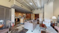 Located at 707 Prudden Street in Lansing, Michigan, this single-floor loft-style unit showcases an industrial-inspired design with soaring ceilings, exposed structural beams, ductwork, and polished concrete floors throughout. The open-concept living area is expansive and airy, anchored by a generously sized seating arrangement that flows naturally into the dining space. The dining area benefits from tall windows and a glass-paned door that invites abundant natural light and provides access to an outdoor area. Large louvered sliding panels add architectural interest and privacy flexibility.

The kitchen features warm maple cabinetry, granite countertops, stainless steel appliances including a refrigerator and gas range, and a central island with bar seating. A set of sliding barn doors and floor-to-ceiling curtain panels serve as stylish room dividers, separating the sleeping areas from the main living space. The primary bedroom is spacious with soaring ceilings and exposed concrete columns, conveying a striking loft aesthetic. Both bathrooms are well-appointed, one featuring a vessel sink and open vanity, and another with a tiled soaking tub and shower combination. A dedicated laundry room with full-size washer and dryer completes the layout.