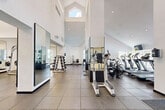 Located at 680 SE 13th St in Dania Beach, Florida, this two-floor fitness facility provides an expansive and well-appointed environment for exercise across both its ground and upper levels. The space is defined by its striking architectural design, most notably the soaring double-height ceilings in the main workout area, which are punctuated by elevated clerestory windows that flood the interior with natural light, creating an open and energizing atmosphere.

The main fitness floor features rubber-tiled flooring throughout, providing durable and functional surfaces suited for a wide range of exercise. Large floor-to-ceiling mirrors are positioned throughout the space, visually expanding the room while supporting training activities. Equipment is thoughtfully arranged and includes cardio machines such as treadmills, ellipticals, stationary bikes, and air bikes, as well as strength-training machines, a cable system, free weights, dumbbells, and a dedicated bench press area.

A separate mirrored studio space with hardwood flooring offers a distinct area for group classes or individual training. The facility also includes restrooms, a water fountain with a bottle-filling station, and clearly marked exits. Windows along the perimeter provide views of surrounding tropical landscaping, reinforcing the bright, welcoming character of this well-equipped fitness center.