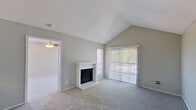 This single-floor residence offers a well-organized layout that balances comfort and functionality across its key living areas. The living room serves as the centerpiece of the home, distinguished by dramatic vaulted ceilings with a textured finish that amplify the sense of space and openness. A white-brick fireplace anchors one wall, while a large sliding glass door fitted with vertical blinds floods the room with natural light and provides direct outdoor access. Soft carpeting throughout adds warmth to the neutral gray palette. Adjacent to the living room, a pass-through window with a granite-style countertop connects to the kitchen, which features dark cabinetry, laminate wood-style flooring, and black appliances including a range, refrigerator, and dishwasher. A secondary room near the entry benefits from a ceiling fan with light fixture and a windowed door that brings in additional natural light. The bathroom is equipped with a full tub-and-shower combination framed by clean white tile, paired with a wide vanity featuring a granite-style countertop, dark cabinetry, and a large mirror. A walk-in closet is accessible directly from the bathroom, offering convenient storage. The home presents a cohesive, understated aesthetic throughout.