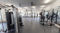 This fitness center is a comprehensively outfitted facility designed to support a full spectrum of workout routines. The space is bright and open, with large windows lining the walls and admitting generous natural light and outdoor views, complemented by modern flush and linear ceiling fixtures. Durable rubber flooring covers the entire area, providing safety and comfort underfoot.

The main workout floor is generously proportioned, housing a robust selection of cardio machines including multiple treadmills and stationary bikes. A dedicated strength training zone features a power rack with weight plates, a Star Trac Instinct functional trainer, and additional cable and resistance machines. A freestanding TRX suspension training rig occupies a central position, offering versatile bodyweight exercise options.

A separate functional training area, distinguished by a bold blue accent wall and glass partition, provides space for dynamic workouts. It is stocked with kettlebells, medicine balls, slam balls, battle ropes, and a wall-mounted television for guided fitness classes. Full-length mirrors are strategically positioned throughout to assist with form and technique.

Ceiling fans ensure consistent air circulation, maintaining a comfortable training environment. The overall design is clean, modern, and organized, creating an inviting and motivating atmosphere for users of all fitness levels.
