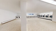 Located at 2233–2235 Hurontario Street in Mississauga, Ontario, this communal laundry facility offers a clean, functional, and spacious environment designed to accommodate high-volume laundry needs. The single-floor space is laid out as one open room, featuring polished terrazzo flooring throughout that is both durable and easy to maintain. Bright white walls and fluorescent strip lighting ensure the entire area is well-illuminated, creating a practical and hygienic atmosphere.

Multiple rows of commercial-grade front-load washing machines and dryers line the perimeter walls, providing ample capacity for residents. The machines are organized in neat, continuous banks, maximizing efficiency and ease of use. A centrally located payment kiosk allows for convenient, cashless transactions. Additional utility features include a utility sink with cabinetry, a folding table for garment handling, and baseboard heaters along the walls for year-round comfort.

Natural light filters in through several windows positioned along the upper portions of the walls, complementing the artificial lighting and giving the space an open, airy feel. Structural columns are integrated into the floor plan without impeding circulation. A clearly marked entry door with safety signage ensures secure and straightforward access. Overall, the facility is thoughtfully organized to deliver a streamlined and comfortable laundry experience.
