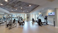 Located at 3220 Hatchet Bay Dr in Charleston, SC, this single-floor fitness center is a standout amenity space defined by both its functional design and distinctive aesthetic character. The most striking architectural feature is the reclaimed wood plank ceiling in the main exercise area, which adds a warm, rustic contrast to the otherwise clean, contemporary interior finished in soft gray tones.

The facility is generously proportioned and divided into clearly defined workout zones. A dedicated cardio section houses a row of treadmills positioned along large windows that offer views of surrounding greenery and palm trees, creating an inviting, distraction-free environment for endurance training. Mounted flat-screen TVs provide additional entertainment throughout the space.

The strength and functional training zones are well-appointed with multiple cable machines, weight benches, a full dumbbell rack, and a variety of selectorized equipment. Additional cardio options include ellipticals, a rowing machine, and a stair climber. Floor-to-ceiling mirrors line several walls, visually expanding the space and supporting proper form during workouts.

Flooring transitions from large-format tile near the entry and cardio zones to a durable rubber-style surface in the strength area. The facility also includes a water fountain station and private restroom access, ensuring a complete and convenient experience. Recessed lighting and ceiling fans throughout maintain a bright, comfortable atmosphere.