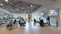 Located at 3220 Hatchet Bay Dr in Charleston, SC, this single-floor fitness center is a standout amenity space defined by both its functional design and distinctive aesthetic character. The most striking architectural feature is the reclaimed wood plank ceiling in the main exercise area, which adds a warm, rustic contrast to the otherwise clean, contemporary interior finished in soft gray tones.

The facility is generously proportioned and divided into clearly defined workout zones. A dedicated cardio section houses a row of treadmills positioned along large windows that offer views of surrounding greenery and palm trees, creating an inviting, distraction-free environment for endurance training. Mounted flat-screen TVs provide additional entertainment throughout the space.

The strength and functional training zones are well-appointed with multiple cable machines, weight benches, a full dumbbell rack, and a variety of selectorized equipment. Additional cardio options include ellipticals, a rowing machine, and a stair climber. Floor-to-ceiling mirrors line several walls, visually expanding the space and supporting proper form during workouts.

Flooring transitions from large-format tile near the entry and cardio zones to a durable rubber-style surface in the strength area. The facility also includes a water fountain station and private restroom access, ensuring a complete and convenient experience. Recessed lighting and ceiling fans throughout maintain a bright, comfortable atmosphere.