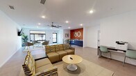 Located at 180 N Martin Luther King Blvd in Lexington, Kentucky, this single-floor community lounge space is designed with a clean, contemporary aesthetic that prioritizes comfort and functionality. The open-plan layout is anchored by a bold burnt-orange accent wall displaying the building's "180" branding in large lettering, serving as a striking focal point throughout the room.

The space is divided into several distinct zones. A central lounge area features a modular sectional sofa with patterned upholstery arranged around a round pedestal coffee table, complemented by additional seating options including accent chairs positioned atop area rugs. A wall-mounted flat-screen television is accessible from multiple seating positions, making the area well-suited for both relaxation and communal viewing.

A dedicated kitchenette is outfitted with navy-blue cabinetry, a stainless steel countertop with sink, and a butcher-block high-top table with bar-height seating — ideal for casual dining or socializing. A workstation area includes a built-in desk and chair alongside a printer, catering to everyday productivity needs.

Natural light enters through large windows near the entrance, where additional casual seating provides a bright spot for conversation. Ceiling fans, recessed lighting, and neutral tile flooring throughout contribute to the space's airy, well-lit atmosphere. Potted plants and framed artwork add warmth and a welcoming finish to the overall environment.
