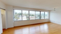 This well-appointed single-floor residence offers a thoughtfully designed open-concept layout that maximizes both space and natural light. The living area is a standout feature, showcasing a dramatic wall of large sliding windows that frame sweeping views of a lush hillside cityscape with palm trees and surrounding neighborhoods, creating a bright and airy atmosphere throughout the day. Warm-toned bamboo hardwood flooring extends across the main living and kitchen areas, lending a clean, modern aesthetic.

The kitchen is efficiently designed with rich, dark walnut-finish cabinetry, crisp white countertops, stainless steel appliances including a range and built-in microwave, and a breakfast bar that opens directly to the main living area — ideal for both daily use and entertaining.

A hallway lined with clean white walls and continued bamboo flooring leads to the private areas of the residence, anchored by a striking set of full-height dark wood built-in cabinetry that provides generous storage. The bedroom features plush grey carpeting and a sliding glass door that opens to a private balcony with additional outdoor views.

The bathroom is finished in a clean, contemporary style with white subway tile surrounding a full soaking tub and shower, hexagonal mosaic floor tile, brushed nickel fixtures, and warm wood vanity cabinetry.