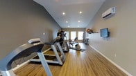 This dedicated fitness space is thoughtfully designed to support a comprehensive workout routine. The room features vaulted ceilings with recessed lighting, creating an open and energizing atmosphere that makes the space feel larger than it is. Warm hardwood-style flooring runs throughout, providing both durability and an inviting aesthetic.

The room is equipped with a solid lineup of professional-grade cardio machines, including a Precor treadmill, an additional treadmill, and a stationary recumbent bike, all positioned to maximize floor space and usability. A multi-function strength training cable machine anchors the workout area, rounding out the equipment offerings for both cardio and resistance training.

Along one wall, a neatly organized rack holds a full set of dumbbells alongside medicine balls, offering versatile options for strength and functional training. A wall-mounted flat-screen television is positioned for easy viewing during workouts. A wall-mounted split air conditioning unit ensures year-round climate comfort. Natural light filters in through a set of windows at the far end of the room, adding brightness and a connection to the outdoors. The neutral gray walls and clean white trim complete the polished, functional design of this well-appointed fitness space.