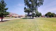 The Woodhaven community park is an inviting outdoor space designed to serve as a central gathering area for the surrounding neighborhood. The grounds are characterized by a generous, well-maintained expanse of open green lawn that provides ample room for recreational activities and leisurely enjoyment. A standout feature of the park is its covered picnic pavilion, which sits beneath the shade of a large, mature tree. The towering tree adds a sense of established character to the space and provides natural shade across a significant portion of the grounds. The pavilion offers a sheltered area ideal for community gatherings, picnics, and outdoor events. Adjacent to the pavilion, a children's playground is equipped with play structures, making the park a family-friendly destination. A paved pathway runs along the perimeter of the space, providing convenient access for pedestrians and cyclists. The park is bordered by the homes of the Woodhaven residential community, creating an integrated neighborhood feel. Under open blue skies, the park presents a bright, cheerful atmosphere that encourages outdoor activity and community interaction year-round.