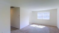 This single-floor space presents a clean and functional layout highlighted by neutral white walls and wall-to-wall carpeting throughout the main living area. The open living space is brightened by a large multi-pane window that allows generous natural light to pour in, casting sunlit patches across the carpeted floor and creating a warm, airy atmosphere. A secondary window is also visible, further enhancing the brightness of the interior. The layout incorporates a partial interior wall that subtly divides the space, adding a degree of architectural definition without fully enclosing separate areas. A thermostat and light switch panel are mounted on the wall, indicating climate control features within the unit. The white ceilings and light-toned walls contribute to an open and uncluttered feel throughout. The carpeting adds a layer of warmth and comfort underfoot, complementing the overall neutral color palette. Electrical outlets are positioned at standard intervals along the baseboards, and a ceiling vent is visible, suggesting a central HVAC system. The space is currently vacant, allowing for a clear view of its structural and design elements.