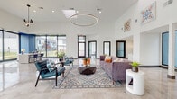 This contemporary single-story facility showcases a thoughtfully designed interior centered around an open, light-filled layout. Polished large-format marble-look tile flooring flows throughout, reflecting natural light and reinforcing the clean, upscale aesthetic. The main lounge area commands attention with soaring ceilings, a striking circular pendant light fixture, and floor-to-ceiling windows that frame outdoor views and flood the space with daylight. Plush seating, a geometric coffee table, and a patterned area rug add warmth and color against the crisp white walls.

Adjacent to the lounge, a kitchenette offers warm wood cabinetry, gray upper shelving with interior lighting, subway tile backsplash, and a beverage refrigerator. A dedicated office area features bold cobalt blue accent walls, large sliding windows overlooking a landscaped exterior, and clean-lined modern furniture. Frosted glass-paneled doors and full-height glass partitions define a private conference room, maintaining visual openness while providing separation.

A well-equipped fitness center rounds out the facility, featuring a distinctive blue and neutral hexagonal patterned floor, multi-function cable machines, free weights, and high ceilings with a bold blue architectural accent. Together, these spaces create a polished, functional, and welcoming environment.