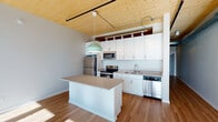 This one-bedroom, one-bathroom residence in Milwaukee's 53204 zip code showcases a thoughtfully designed modern interior distinguished by striking exposed wood plank ceilings that lend warmth and an industrial-contemporary character throughout the open layout. Rich hardwood flooring runs continuously across the single-floor plan, complementing the clean architectural aesthetic.

The kitchen serves as a focal point of the living space, featuring white shaker-style cabinetry extending to the ceiling, a full suite of stainless steel appliances including a refrigerator, electric range, over-the-range microwave, and dishwasher, along with a central island with a neutral countertop that provides additional workspace and casual seating potential. A mint-toned pendant light adds a subtle design accent above the island.

The bedroom is generously filled with natural light courtesy of tall, floor-to-ceiling windows framed in sleek black trim, offering open views of the Milwaukee skyline and surrounding cityscape. Exposed ductwork and track lighting integrated into the wood ceiling reinforce the unit's modern industrial style.

The bathroom is finished with a walk-in shower unit and a built-in tall storage cabinet, providing practical organization. Overall, the unit presents a cohesive, contemporary aesthetic with quality finishes throughout.
