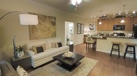 This single-story space showcases an open-concept layout that connects the living, dining, and kitchen areas into one cohesive, flowing environment. The living area features vaulted ceilings that enhance the sense of spaciousness, complemented by warm hardwood-style flooring that extends throughout. A large, decorative canvas with gold and earth tones anchors the living space, which is furnished with a cream sofa, patterned area rug, and a dark espresso coffee table. Overhead, a multi-light ceiling fixture provides ambient illumination. Adjacent to the living area, a dining nook accommodates a dark wood table with upholstered high-back chairs, situated beneath elegant pendant lighting. The kitchen, visible from the main living space, features warm wood cabinetry, dark countertops, and track lighting, contributing to the overall cohesive design. A doorway in the background reveals a bright secondary room, suggesting additional living space beyond. The neutral warm-toned walls and layered lighting throughout create an inviting, well-appointed ambiance suitable for both everyday living and entertaining.