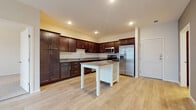 This single-floor residence offers a thoughtfully designed layout that balances functionality and modern aesthetics throughout. The kitchen serves as the centerpiece of the home, showcasing rich dark espresso-toned cabinetry that extends to the ceiling, granite countertops, and a full suite of stainless steel appliances including a French door refrigerator, range, built-in microwave, and dishwasher. A freestanding kitchen island with a matching granite top and open shelving provides additional prep space and storage, all set atop light-toned hardwood-style flooring that extends throughout the main living areas. Recessed lighting illuminates the kitchen and entry, creating a bright and welcoming atmosphere. The bedrooms are finished with plush neutral carpeting and feature large windows fitted with horizontal blinds, allowing for ample natural light and views of the surrounding neighborhood. Baseboard heating units run beneath the windows, ensuring consistent comfort. The bathroom is elegantly appointed with dark wood vanity cabinetry, granite countertops, a framed mirror, and a tall linen tower cabinet for additional storage. Light wood-style flooring continues into the bathroom, complementing the neutral wall tones. A dedicated in-unit laundry area houses a full-size top-loading washer and dryer with overhead wire shelving, adding everyday convenience to this well-rounded residence.
