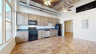 This single-floor unit showcases an industrial-loft aesthetic with thoughtfully modern finishes throughout. The open living area immediately impresses with its soaring ceilings, exposed metal ductwork, and ceiling fans that complement the raw architectural character of the space. Warm, light-toned hardwood flooring extends throughout the main living areas, enhancing the sense of openness and continuity.

Large, floor-to-ceiling windows at the front of the unit allow abundant natural light to pour in while offering views of the surrounding streetscape. An original exposed brick column adds a distinctive architectural detail, reinforcing the building's historic character. The entry area features a classic paneled door framed by transom windows, further amplifying the light-filled atmosphere.

The kitchen is well-appointed with light natural wood shaker-style cabinetry extending to the ceiling, dark countertops, and a full suite of appliances including an electric range, built-in microwave, dishwasher, and refrigerator. Hardwood flooring continues seamlessly into this space.

The bathroom features tile flooring, a tub-shower combination with floor-to-ceiling white tile surround, dark wood vanity cabinetry, and a large mirror. A dedicated laundry area with washer/dryer hookups, upper cabinetry, and a pet door adds convenient functionality to the unit.
