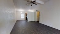 This single-floor space is defined by its open, airy layout and thoughtful design details that create a welcoming and functional environment. The main living area immediately draws the eye upward with its vaulted, beamed ceiling, which adds a sense of height and volume to the room. A ceiling fan with a light fixture is centrally mounted, providing both comfort and illumination. The dark hardwood flooring runs throughout, offering a rich contrast against the crisp white walls and lending the space a grounded, contemporary feel. Natural light enters through a large window dressed with vertical blinds, brightening the open floor plan. A partial room divider wall separates the living area from the kitchen zone, maintaining an open connection while providing subtle definition between spaces. The kitchen area features wood cabinetry and a stainless steel refrigerator, visible from the main living space. An elegant chandelier marks the dining area, adding a touch of refinement. A hallway leads to additional rooms, with louvered closet doors visible, hinting at practical storage solutions throughout. The overall ambiance is clean, modern, and move-in ready, with neutral tones and architectural details that provide a versatile canvas for any interior style.