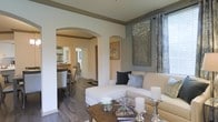 This single-floor residence showcases a thoughtfully designed interior that balances elegance with everyday comfort. The living area serves as the heart of the home, featuring rich hardwood flooring, neutral-toned walls accented with warm gray crown molding, and large windows dressed with layered curtains that invite natural light into the space. A plush sectional sofa anchors the room, complemented by a rustic wooden coffee table adorned with decorative candle holders and fresh florals, creating an inviting and refined atmosphere.

Distinctive arched doorways define the architectural character of the space, providing graceful transitions between rooms while maintaining an open, connected feel. Through the arches, the dining area is visible, complete with a dark wood dining table and upholstered chairs, and it flows seamlessly toward the kitchen beyond, which is illuminated with bright lighting. Wall art and carefully chosen décor accents throughout the living space add personality and warmth without overwhelming the clean, cohesive aesthetic.

The overall ambiance is sophisticated yet approachable, with a neutral color palette, classic architectural details, and curated furnishings that together create a welcoming and well-appointed home environment.