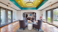 The Wimbledon Apartments leasing office is a single-floor community space designed to make a bold and welcoming first impression. The centerpiece of the space is the striking main lobby, where a raised tray ceiling painted in vivid yellow and teal immediately draws the eye upward, complemented by a statement chandelier with exposed Edison-style bulbs. Warm terra cotta tile flooring runs throughout, adding a grounded, earthy contrast to the energetic color palette overhead.

The seating area is thoughtfully arranged with four plush grey wingback chairs surrounding a round wood-and-metal coffee table, all set atop a plush area rug. Large glass double doors along the far wall flood the room with natural light while offering direct views of the outdoor pool area. An arched passageway leads to a secondary lounge and service area, which features a dedicated coffee bar with white cabinetry, a built-in mini-fridge, and vibrant abstract artwork displayed against a textured grey wood-panel accent wall.

A branded reception area displaying the Wimbledon Apartments logo serves as a focal point for resident and guest check-ins, flanked by lush greenery and additional seating. A wall-mounted display screen and ample counter space round out the functional amenities. Together, these elements create a community hub that is both visually dynamic and practically designed.