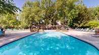 Located at 1630 Crossings Pkwy in Westlake, Ohio, this two-floor property offers an impressive outdoor amenity complex set against a backdrop of mature trees and well-maintained greenery. The centerpiece of the outdoor area is a generously sized swimming pool with sparkling blue water, surrounded by an expansive concrete deck lined with teal and blue lounge chairs ideal for sunbathing. Brick-accented raised planters with colorful seasonal flowers add decorative charm throughout the pool surround.

Adjacent to the pool, a paver-stone patio provides a refined outdoor dining area, complete with shaded seating beneath striped market umbrellas and views of a serene hot tub nestled within the landscaping. A pergola structure with additional seating anchors one end of the pool area, offering a shaded retreat.

The clubhouse building features a distinctive A-frame roofline with large windows and a covered porch, accessible via a broad staircase flanked by brick pillars adorned with potted plants. The grounds also include a dedicated green space with a winding concrete walking path shaded by tall trees, enclosed by ornamental iron fencing. The overall ambiance is resort-like, combining thoughtful landscaping with well-appointed outdoor leisure spaces.