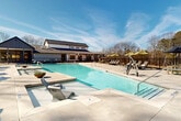 This outdoor aquatic facility centers around a generously sized rectangular swimming pool with crystal-clear turquoise water, accessible via multiple stainless steel entry rails and equipped with an accessibility lift. The surrounding concrete pool deck is expansive, providing ample space for lounging and circulation. The deck is well-furnished with modern chaise loungers and seating clusters shaded by yellow-green market umbrellas, offering both sun and shade options for guests.

A covered open-air pavilion anchors one side of the facility, providing additional shaded seating and a gathering space adjacent to the pool. Nearby, a compact utility structure serves the pool's operational needs. The entire pool area is enclosed by a black metal perimeter fence, ensuring safety while preserving sightlines to the surrounding natural landscape of tall pine and deciduous trees.

The two-story clubhouse building, finished in a modern farmhouse style with board-and-batten siding and a dark pitched roof, overlooks the pool area and features large windows that bring in abundant natural light. The overall ambiance is open, well-maintained, and resort-like, seamlessly integrating designed outdoor amenities with a serene wooded setting.