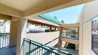 This exterior walkway and balcony space is part of a multi-story building complex characterized by a consistent architectural style. The structure features textured stucco walls finished in a warm beige tone, complemented by green metal railings that line the open-air corridors and balconies throughout the building. The walkways are constructed of smooth concrete and provide access along the building's exterior, offering open views of the surrounding courtyard and neighboring building sections below.

The green accents extend to a prominent canopy or awning visible overhead, adding a pop of color against the clear blue sky and tying together the building's cohesive color scheme. The multi-level design of the complex is evident from the balcony vantage point, where stacked corridors and repeating columns create a structured, layered appearance.

Mature trees and landscaping are visible within the courtyard below, providing natural greenery that softens the hardscape elements of the building. The open design of the walkways allows for abundant natural light and fresh air, with unobstructed sightlines across the complex. The overall ambiance is bright, airy, and well-maintained, reflecting a functional yet visually unified architectural approach suited to a warm, sunny climate.