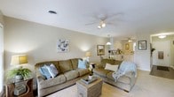 This single-floor residence offers a welcoming and well-designed layout with a relaxed coastal aesthetic throughout. The living area serves as the heart of the home, featuring plush, earth-toned sofas accented with soft blue and neutral throw pillows that complement the light, airy color palette of the space. A wicker coffee table adds a natural, textural element to the seating arrangement, reinforcing the casual yet refined coastal theme. A ceiling fan with integrated lighting overhead provides both comfort and illumination, while warm accent lamps contribute to a cozy ambiance. Coastal-inspired wall art adds thoughtful decorative touches. The open-concept design allows the living area to flow naturally into the kitchen, which is visible in the background and features white cabinetry, light countertops, and pendant lighting. The kitchen appears bright and functional, with clean lines that maintain the overall lightness of the space. Neutral-toned walls and light flooring carry throughout, creating a cohesive and calming atmosphere. The single-floor layout ensures easy accessibility and an uninterrupted sense of openness, making this space both practical and inviting for everyday living.