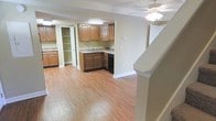 This two-story space offers a functional and comfortable layout with notable features throughout. The kitchen is a central highlight, equipped with warm-toned wood cabinetry that extends to the upper walls, providing ample storage. Granite-style countertops add a polished finish, while a black appliance suite contributes a clean, contrasting aesthetic. An open built-in shelving unit near a doorway offers additional display or storage options, enhancing the kitchen's practicality.

The flooring throughout the main level consists of hardwood-style planks in a natural honey tone, lending warmth and visual continuity to the space. The walls are finished in a neutral beige palette, creating a bright and inviting atmosphere. A ceiling fan with a light fixture provides both air circulation and ambient lighting overhead.

A staircase is visible in the foreground, connecting the two levels of the space, with clean white trim and a simple newel post adding a classic architectural touch. The open-concept feel between the kitchen and the adjacent living area allows for easy flow and versatile use of the space, making it well-suited for everyday living.