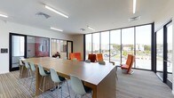 This contemporary single-floor workspace is designed to foster collaboration and productivity in a bright, open environment. The space is anchored by a large, L-shaped wooden work table surrounded by modern light gray chairs, providing ample room for group work or individual tasks. Complementing the workstations, a lounge area features bold orange high-back chairs and a sleek sofa, offering a more relaxed setting for informal meetings or breaks.

Floor-to-ceiling windows with slim black frames run along an entire wall, flooding the interior with abundant natural light and offering expansive views of the surrounding campus or urban landscape. The white ceiling is equipped with recessed linear LED lighting, ensuring consistent illumination throughout. Patterned carpet tiles in neutral tones provide a professional yet comfortable underfoot experience.

An enclosed glass-walled room is visible in the background, likely serving as a private office or meeting room, adding functional versatility to the layout. The overall design palette — clean whites, warm wood tones, and vibrant orange accents — creates a dynamic, energizing atmosphere well-suited to a modern educational, corporate, or co-working environment.
