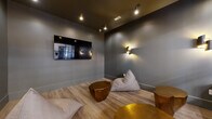 Located at 4275 Valley St in Enola, PA, this compact yet thoughtfully designed lounge room offers a sophisticated and inviting atmosphere. The space is defined by deep charcoal-gray walls and ceiling, which establish a moody, intimate ambiance throughout. Light wood-plank flooring provides a warm contrast to the dark palette, grounding the room with a natural, earthy element.

Lighting plays a central role in the room's character. Sculptural gold-finish wall sconces cast a warm, directional glow across the walls, complementing a set of exposed Edison-style pendant and track lights overhead. A large wall-mounted flat-screen television anchors one wall, making the space well-suited for media viewing and social gathering.

Seating is casual and inviting, featuring a sleek black leather sofa, oversized floor-cushion bean bags in a neutral taupe, and an assortment of distinctive geometric gold-finished side and coffee tables that serve as striking decorative focal points. The mix of seating styles encourages relaxed, informal use of the space.

A full-length glass partition wall with black framing provides visual connectivity to an adjacent, brightly lit common area, allowing natural light to filter in while maintaining a sense of enclosure. A glass entry door reinforces this transparency. The overall design balances modern industrial aesthetics with cozy, lounge-style comfort.