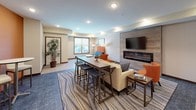This well-appointed community lounge space is thoughtfully designed to serve as a versatile gathering area for residents and guests alike. The open, single-floor layout flows seamlessly between distinct functional zones, creating a cohesive yet dynamic environment.

The kitchenette area is a standout feature, showcasing rich dark cabinetry paired with sleek granite countertops and a striking blue hexagonal tile backsplash that adds a bold, contemporary accent. Stainless steel appliances, including a refrigerator and built-in microwave, complement the space, while a deep undermount sink and generous counter space make it well-suited for entertaining. A high-top bar counter with seating provides an informal dining option adjacent to the kitchenette.

The lounge area centers around a modern linear fireplace set against a wood-paneled accent wall, with a wall-mounted flat-screen television above. Comfortable seating, including a cream sofa and vibrant orange accent chairs, is arranged on a patterned area rug to create an inviting conversation nook. A long communal high-top table with bar stools offers additional flexible seating for work or socializing.

Warm orange accent walls punctuate the space alongside neutral tones, recessed lighting illuminates the interior evenly, and large windows invite natural light into the lounge area, completing this modern and welcoming communal retreat.