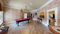 Located at 325 Spears Creek Church Rd in Columbia, SC, this single-floor recreation and game room is a well-appointed communal space designed for leisure and social interaction. The room is defined by its striking exposed brick walls, which lend a warm, rustic character throughout, complemented by crown molding and recessed lighting that keep the space bright and inviting.

The hardwood-style flooring anchors the room and flows seamlessly across its generous open layout. A centerpiece billiards table with a vivid red felt surface is positioned under a classic Craftsman-style pendant light, creating a natural focal point. Adjacent to it, a foosball table adds to the game-room atmosphere. Multiple seating arrangements — including pub-height tables with stools and standard dining-height tables with chairs — offer versatile spaces for casual conversation or tabletop games.

A wall-mounted flat-screen television is positioned for easy viewing from the seating area, while a built-in service counter with cabinetry provides a convenient refreshment area. Framed artwork adorns the brick walls throughout, adding a curated aesthetic touch.

French doors with decorative fabric valances open directly to an outdoor pool area, flooding the room with natural light and providing seamless indoor-outdoor connectivity. This recreation room serves as a vibrant hub for community engagement and entertainment.