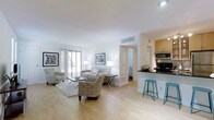 This single-floor space offers a clean, modern aesthetic defined by an open-concept layout that effortlessly connects the living area and kitchen. Light hardwood flooring runs throughout, lending warmth and continuity to the space. The living area is generously proportioned, centered around a comfortable seating arrangement with patterned accent chairs, a sofa, and a glass coffee table resting on a soft area rug. A large sliding glass door floods the room with natural light and provides a view of lush greenery outside, enhancing the bright, airy ambiance. Crisp white walls and a smooth white ceiling with recessed lighting contribute to the fresh, open feel. Tasteful framed artwork adorns the walls, adding character without overwhelming the minimalist design. The kitchen features light wood cabinetry, dark countertops, and a peninsula with a black surface that doubles as a casual dining or breakfast bar, accompanied by two teal metal stools that introduce a subtle pop of color. Modern stainless steel appliances, including a microwave, are neatly integrated into the kitchen space. A doorway visible from the main living area leads to additional rooms, suggesting a thoughtfully connected and functional floor plan throughout.