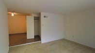 This single-floor residential space presents a clean, functional layout designed for comfortable everyday living. The interior features a combination of soft carpeting in the main living area and hardwood-style flooring in the adjoining room, creating a natural visual distinction between spaces. Neutral white walls throughout contribute to a bright, open atmosphere that allows for versatile interior styling.

A notable feature of the space is the built-in closet, which includes a dedicated shelving unit above the hanging rod, offering practical and organized storage. An HVAC vent is integrated into the wall, ensuring climate comfort year-round. The transition between the carpeted and hard-floored areas is clearly defined, suggesting a thoughtful separation of functional zones within the home.

The space is bathed in warm, ambient light, accentuated by the neutral tones of the walls and ceiling. The overall layout is straightforward and efficient, making it well-suited for those seeking a no-fuss, move-in ready environment. The single-floor design ensures easy accessibility throughout, with a cohesive flow from one area to the next.