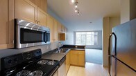 This single-floor residence offers an efficient, open-concept layout that seamlessly connects the kitchen and living areas. The kitchen is well-equipped with light maple cabinetry providing ample storage, dark countertops, and stainless steel appliances including an electric range, overhead microwave, and a double-basin sink. Track lighting above the kitchen workspace ensures functional illumination for meal preparation. The light hardwood flooring in the kitchen transitions naturally into the living area, which features plush carpeting underfoot. A large picture window anchors the living space, flooding the interior with natural light and offering views of surrounding greenery. The soft blue-gray walls throughout create a calm, cohesive atmosphere. The open flow between the kitchen and living area enhances the sense of space, making the layout feel practical and inviting. This residence is well-suited for those seeking a straightforward, functional living space with a clean, modern aesthetic.