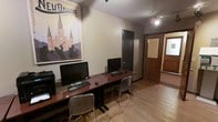 This single-floor space is designed as a functional computer workstation area, offering a practical and organized environment for productivity. The room features light wood laminate flooring that adds a warm tone to the otherwise utilitarian layout. A long, rich mahogany-toned desk runs along one wall, accommodating multiple desktop computer setups complete with monitors, keyboards, and mice — making the space well-suited for shared or individual computer use. Acrylic and metal chairs provide seating at the workstations, offering a modern, lightweight aesthetic. The walls are painted in a neutral beige tone, contributing to a calm and focused atmosphere. A prominent New Orleans travel poster — boldly declaring "The Big Easy" and depicting the iconic Jackson Square cathedral skyline — serves as a decorative focal point, adding personality and a touch of local culture to the space. A dark storage locker or cabinet stands against the back wall, providing additional utility. Natural and artificial lighting illuminate the area, with a ceiling light fixture visible near the entryway. A wood-paneled door leads to an adjacent corridor or room, suggesting the space is part of a larger facility. The textured ceiling completes the interior, giving the room a grounded, practical character throughout.