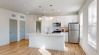 This well-designed 1-bedroom, 1-bathroom unit offers a clean, contemporary aesthetic throughout its efficiently organized single-floor layout. The open-concept living and kitchen area serves as the heart of the space, anchored by a large central island with a crisp white quartz countertop and an integrated sink. The kitchen is outfitted with full stainless steel appliances, including a refrigerator, electric range, and over-the-range microwave, complemented by white shaker-style cabinetry, a classic subway tile backsplash, and globe pendant lights that add a modern touch. Light hardwood-style flooring runs continuously throughout the unit, enhancing the bright and airy atmosphere created by recessed lighting and multiple windows. The spacious living area benefits from a ceiling fan and abundant natural light filtering through slatted blinds. The bedroom is a comfortable, well-proportioned retreat, also featuring a ceiling fan and a natural light-filled window. A dedicated walk-in closet with wire shelving provides generous storage. The bathroom maintains the unit's clean design language with white cabinetry, a quartz vanity top, a large mirror, and a tiled shower-tub combination, all set on matching hardwood-style flooring.