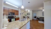 This thoughtfully designed 2-bedroom, 1-bathroom residence offers a comfortable and functional single-floor layout. The open-concept living area features warm hardwood flooring, crown molding, and a welcoming living room with natural light filtering through a large window. The kitchen is a standout space, outfitted with rich cherry wood cabinetry, granite countertops, black appliances including an electric range and built-in microwave, a double-basin sink set in a peninsula island, and pendant lighting overhead. A dedicated dining area adjacent to the kitchen accommodates a four-seat table and is accented by track lighting and crown molding details. Both bedrooms are carpeted and equipped with ceiling fans, offering a quiet retreat. The primary bedroom is generously sized with ample natural light from large windows and access to a spacious walk-in closet featuring wire shelving. The full bathroom includes a wood-toned vanity with a granite-style countertop, a soaking tub with shower combination, and hardwood-style flooring. An additional closet provides extra storage. The overall ambiance is clean, neutral, and move-in ready, with consistent finishes throughout that create a cohesive and practical living environment.
