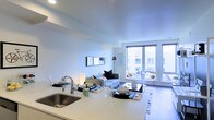 This single-floor space showcases a contemporary open-concept layout that seamlessly connects the kitchen and living areas into one cohesive, functional environment. The kitchen features light-toned countertops, a stainless steel sink, and a clean, minimalist aesthetic that transitions naturally into the dining and living zones. The living area is bright and airy, benefiting from large floor-to-ceiling windows that flood the space with natural light and frame views of the surrounding urban landscape. Sliding glass doors lead to a private balcony, extending the living space outdoors and offering additional exposure to natural light. The flooring throughout is hardwood, adding warmth and continuity across the open layout. Recessed lighting in the ceiling complements the abundant natural light, ensuring the space remains well-illuminated at all times. The overall design leans toward a modern, neutral palette — white walls, light counters, and natural wood tones — creating a versatile and inviting atmosphere. The thoughtful layout maximizes the sense of openness while maintaining clearly defined functional zones within the shared living space.