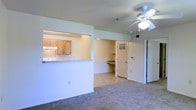 This single-floor residence is designed with an open and functional layout that maximizes the use of space. The living area features neutral-toned carpeting, which lends a warm and understated character to the room. A ceiling fan with an integrated light fixture is centrally mounted, providing both ventilation and ambient lighting.

The kitchen is positioned adjacent to the living space and is accessible via a pass-through counter, which creates a sense of connectivity between the two areas while maintaining a defined boundary. The kitchen is equipped with light wood cabinetry and a white countertop, offering ample storage and preparation space. An overhead exhaust range hood is visible above the cooking area.

Beyond the living room, a hallway leads to additional areas of the unit. Notable is a built-in desk or shelving nook, providing a dedicated space for work or storage. A door with a louvered window panel adds architectural detail while allowing light to filter through. The overall color palette is neutral and bright, with white walls throughout, contributing to an airy and clean aesthetic. The layout is practical and well-organized.