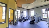 Located at 75 Cross Creek Pkwy in Hattiesburg, Mississippi, this single-floor fitness center is designed to provide a well-equipped and inviting workout environment. The space features a modern aesthetic, highlighted by a bold color scheme of warm yellow and cool gray tones, accented by a distinctive tray ceiling with a warm-toned inset that adds architectural interest and depth to the room.

Natural light floods the interior through multiple large, grid-pane windows positioned along the walls, creating a bright and energizing atmosphere. A ceiling fan is centrally mounted beneath the tray ceiling, ensuring comfortable airflow throughout workout sessions. A wall-mounted television is also present, offering entertainment during exercise.

The fitness floor is finished with durable speckled rubber flooring, ideal for high-traffic athletic use. The equipment lineup includes elliptical machines, stationary bikes, treadmills, and strength training machines, providing a comprehensive range of cardio and resistance options for users of all fitness levels.

Glass-panel doors open into an adjacent lounge or breakout area, visible through the transparent partition and featuring a cheerful yellow accent wall, adding a sense of connectivity and openness to the overall layout. The combination of thoughtful design and functional equipment makes this facility a well-rounded, comfortable space for fitness activity.