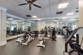 This fitness center offers a generously sized, single-level workout space designed to accommodate a wide range of exercise needs. The layout is thoughtfully organized, with distinct zones dedicated to strength training, free weights, and functional fitness equipment. Multiple selectorized cable and resistance machines are positioned throughout the open floor plan, complemented by a full dumbbell rack, adjustable benches, and functional training accessories such as medicine balls and stability balls.

The interior design reflects a refined aesthetic, featuring white-painted columns, crown molding, and neutral walls that lend the space an upscale feel uncommon in typical fitness facilities. A drop ceiling with recessed panel lighting and multiple ceiling fans provides consistent, even illumination while maintaining comfortable air circulation throughout. Large windows along multiple walls invite abundant natural light and offer views of the surrounding outdoor greenery, creating an airy, open atmosphere.

Floor-to-ceiling mirrors line key sections of the walls, visually expanding the space and providing practical feedback during exercise. The durable speckled rubber flooring offers safety and stability across the entire workout area. A carpeted entryway transitions cleanly into the main fitness floor, and multiple exit doors provide convenient access points throughout.