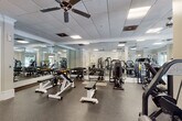 This fitness center offers a generously sized, single-level workout space designed to accommodate a wide range of exercise needs. The layout is thoughtfully organized, with distinct zones dedicated to strength training, free weights, and functional fitness equipment. Multiple selectorized cable and resistance machines are positioned throughout the open floor plan, complemented by a full dumbbell rack, adjustable benches, and functional training accessories such as medicine balls and stability balls.

The interior design reflects a refined aesthetic, featuring white-painted columns, crown molding, and neutral walls that lend the space an upscale feel uncommon in typical fitness facilities. A drop ceiling with recessed panel lighting and multiple ceiling fans provides consistent, even illumination while maintaining comfortable air circulation throughout. Large windows along multiple walls invite abundant natural light and offer views of the surrounding outdoor greenery, creating an airy, open atmosphere.

Floor-to-ceiling mirrors line key sections of the walls, visually expanding the space and providing practical feedback during exercise. The durable speckled rubber flooring offers safety and stability across the entire workout area. A carpeted entryway transitions cleanly into the main fitness floor, and multiple exit doors provide convenient access points throughout.