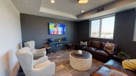This contemporary single-floor communal space is thoughtfully designed to accommodate both relaxed socializing and lively gatherings. The layout flows seamlessly between distinct functional zones, each finished in a cohesive, modern palette of deep charcoal and neutral tones.

The lounge area is anchored by a large wall-mounted flat-screen TV set against a bold dark accent wall, complemented by a rich brown leather sectional sofa, plush upholstered armchairs, and a distinctive cylindrical rattan coffee table. A tray ceiling with recessed and pendant lighting adds architectural depth, while a window offers views of the surrounding cityscape.

Adjacent to the lounge, a spacious seating nook features a pair of elegant wingback chairs arranged around a compact side table beneath a raised tray ceiling, creating an intimate setting for conversation.

The dining and bar area is a standout feature, showcasing expansive reclaimed-wood bar-height tables lined with padded round stools. Large windows along this zone flood the space with natural light and frame sweeping outdoor views, enhancing the open, elevated ambiance.

The kitchen is outfitted with stainless steel appliances, including a gas range with a sleek overhead hood, a full-size refrigerator, and dark countertops, all set against large-format tile flooring and a bright window overlooking the city.
