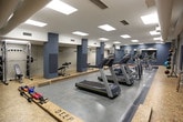 Located at 130 Brooke Ave in Norfolk, Virginia, this fitness center offers a well-equipped, single-floor exercise facility designed to support a comprehensive workout experience. The space features a generous open floor plan with a smooth, durable concrete floor in the main exercise area, complemented by a raised cork or rubberized platform along the perimeter — ideal for stretching, free weight exercises, and functional training. Structural columns finished in a deep slate blue add a modern industrial character throughout the room. Overhead, a grid of recessed panel lighting provides bright, even illumination across the entire facility. The cardio zone is prominently featured, housing a row of treadmills, stationary bikes, and elliptical machines arranged for easy access and circulation. A dedicated strength training section includes weight benches, cable machines, and a full rack of dumbbells. Large wall-mounted mirrors expand the visual depth of the space and allow users to monitor their form. Additional amenities include wall-mounted accessory storage with resistance bands and medicine balls, stability balls, and mounted flat-screen televisions. A utility area features stainless steel water fountains and a waste receptacle set against tile-clad walls, ensuring convenience and hygiene throughout the facility.