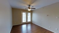 This single-floor space presents a clean, functional layout with notable design details that contribute to its overall appeal. The main living area is anchored by rich, dark hardwood floors that run throughout, reflecting natural light and adding warmth to the room. A classic five-blade ceiling fan with an integrated light fixture is centrally mounted, providing both air circulation and ambient lighting.

A standout feature of this space is the set of French doors, which serve as the primary source of natural light. The glass-paneled doors allow sunlight to flow generously into the interior while offering direct access to an outdoor area, creating a seamless indoor-outdoor connection. The neutral, beige-toned walls provide a versatile backdrop that complements the flooring and keeps the space feeling open and airy.

White baseboards trim the perimeter of the room, adding a crisp contrast to the warm flooring and walls. The overall ambiance is bright, tidy, and welcoming, with a straightforward design that emphasizes practicality while maintaining a comfortable aesthetic suitable for a variety of uses.