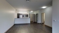 This single-floor space features an open-concept design that seamlessly integrates the living area and kitchen into one cohesive environment. The kitchen is outfitted with dark cabinetry, stainless steel appliances including a refrigerator and built-in microwave, and a light-toned granite or stone countertop that extends into a peninsula-style breakfast bar, providing both prep space and a casual dining option. Recessed and flush-mount ceiling lighting, along with a linear pendant fixture above the kitchen area, ensures consistent illumination throughout. The flooring consists of dark hardwood-style planks that run continuously across the open living space, adding warmth and visual continuity. Walls are finished in a clean, neutral white, contributing to a bright and modern aesthetic. Toward the rear of the unit, multiple doorways lead to additional rooms, with one appearing to offer a glimpse of a bathroom. The layout is practical and well-organized, with clear sightlines across the main living area. Overall, the space presents a contemporary, functional design with clean finishes and a straightforward, efficient floor plan.