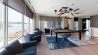 Located at 25 Lewis St in Boston's East Boston neighborhood, this single-floor communal lounge is a thoughtfully designed social space that blends contemporary aesthetics with functional comfort. The open-plan layout is anchored by wide-plank hardwood flooring that flows seamlessly throughout, tying together distinct zones within the space.

The main lounge area features a generous sectional sofa in soft gray tones, accented with yellow throw pillows and complemented by a bold, abstract area rug in deep blues and charcoal. A striking yellow accent chair adds a pop of color, while warm wood wall paneling with integrated ambient lighting creates a sophisticated, layered backdrop.

Deeper into the space, a games area centers around a premium billiards table set atop another dramatic patterned rug. Overhead, a sculptural chandelier with angular black and gold elements serves as a visual focal point. Navy lounge chairs flank the perimeter, and large floor-to-ceiling windows with sheer drapery flood the room with natural light while framing captivating waterfront views of the Boston Harbor.

A bar or service counter with white tile detailing is visible in the background, adding to the versatility of the space. Teal accent walls and lush indoor greenery provide vibrant contrast throughout, completing the polished, resort-inspired atmosphere of this exceptional amenity space.
