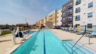 This outdoor pool area is a well-appointed amenity space designed for both recreation and relaxation. The centerpiece is a generously sized swimming pool with clear blue water, framed by a wide concrete deck lined with lounge chairs ideal for sunbathing. An ADA-compliant pool lift provides accessible entry into the water, reflecting a thoughtful, inclusive design approach.

Surrounding the pool, the architecture features a mix of natural stone masonry, warm wood accents, and contemporary siding in contrasting tones of yellow, brown, and gray. A covered outdoor corridor runs along the building's ground level, supported by substantial stacked-stone columns with warm wood beam detailing overhead. This shaded walkway provides a comfortable transition between the pool area and interior access points, and is furnished with cushioned seating for additional lounging.

The pool area is enclosed by a black iron security fence with a key-coded entry gate, ensuring privacy and controlled access. Exterior wall-mounted lighting along the covered walkway adds functionality after dark. Views of the surrounding grounds, including open green space and the multi-story building facade with private balconies, create an inviting courtyard atmosphere. The overall ambiance is clean, modern, and resort-inspired, offering a compelling outdoor retreat.