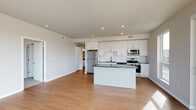 Located at 1501 NW 38th St in Kansas City, Missouri, this modern single-floor unit offers a clean, contemporary design throughout. The open-concept living and kitchen area is anchored by warm wood-tone flooring and recessed lighting. The kitchen is well-appointed with white shaker-style cabinetry, dark granite countertops, a white tile backsplash, a central island, and a full suite of stainless steel appliances including a refrigerator, electric range, built-in microwave, and dishwasher. The living area is bathed in natural light through an expansive bank of large windows and offers direct access to a private balcony overlooking the surrounding landscape. The bedrooms feature plush carpeting and generous closet space, with large windows that frame outdoor views and invite ample daylight. The bathroom is finished with a white vanity, dark granite countertop, undermount sink, and a large mirror, maintaining the consistent modern aesthetic found throughout the unit. Neutral tones, clean lines, and quality finishes define the overall ambiance, creating a bright and functional living environment.